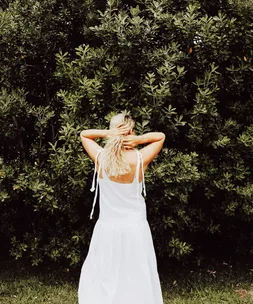 Back view of a woman in a white dress holding her hair, standing in front of lush green bushes.