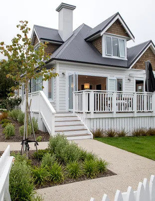 White house with gray roof, front porch, and garden path surrounded by greenery and small tree.