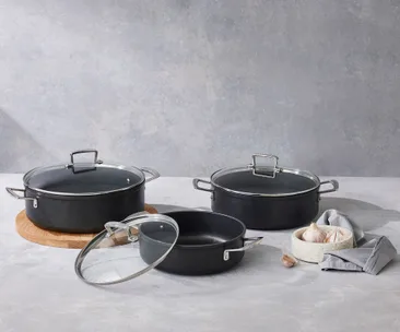 Cookware set with glass lids on a countertop, accompanied by garlic bulbs in a bowl and a grey cloth nearby.