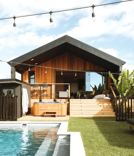 Contemporary wooden house with a pool, hot tub, and patio, accented with string lights and lush greenery.