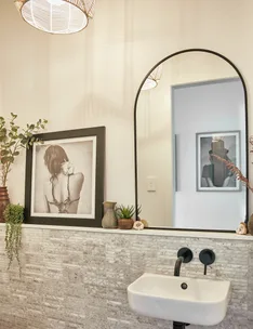 Modern bathroom with a framed photo, mirror, plants, and a white washbasin against textured tile wall.