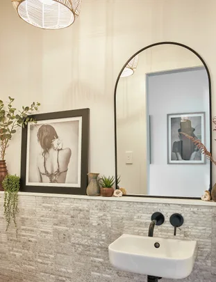 Modern bathroom with a framed photo, mirror, plants, and a white washbasin against textured tile wall.