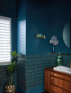 A bathroom with dark blue walls, green plants, a small painting, a round mirror, and a wooden sink cabinet.