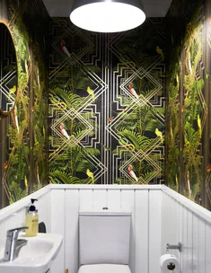 A small bathroom with a vibrant bird and leaf-patterned wallpaper, white paneling, and a round mirror above a sink.