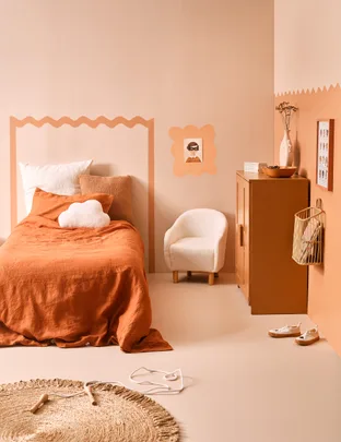 Cozy bedroom with orange bedding, white cloud pillow, round rug, beige chair, and minimal decor.