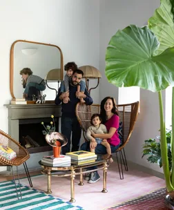 A family of four in a modern living room with large plants and colorful decor. The father holds one child on his shoulders.