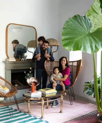 A family of four in a modern living room with large plants and colorful decor. The father holds one child on his shoulders.