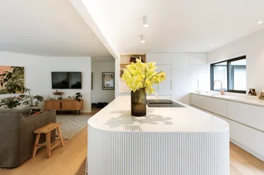 Modern kitchen with a white island countertop, yellow flowers in a vase, and a living area with TV and plants.