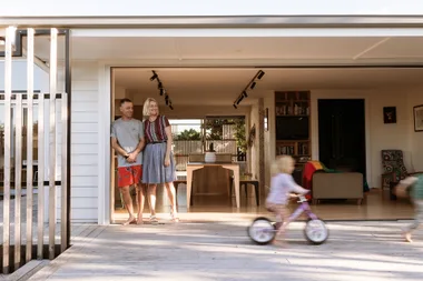 A couple smiles at their home's open glass door while a child rides a bike on the patio.