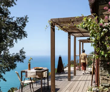Person walking on a wooden deck overlooking the sea, surrounded by pergolas and greenery.