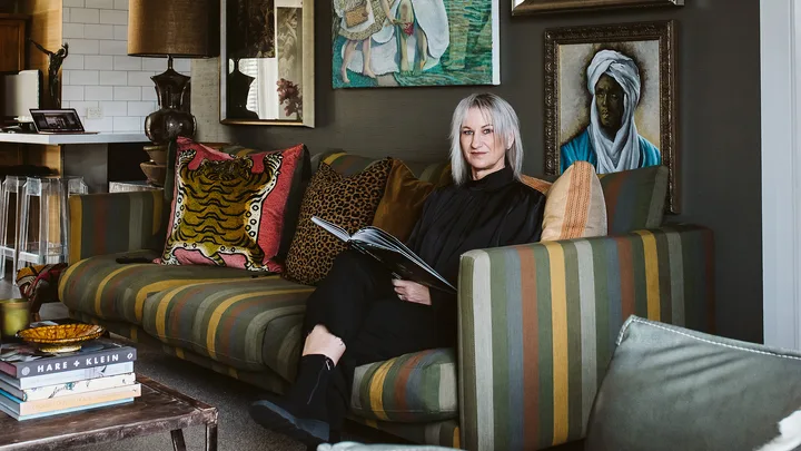 A woman sitting on a striped sofa with colorful cushions in a room with framed artwork and a table with stacked books.