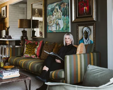 A woman sitting on a striped sofa with colorful cushions in a room with framed artwork and a table with stacked books.
