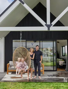 Family standing on a porch; two children sit in a hanging chair, and two adults stand beside them in front of a modern house.