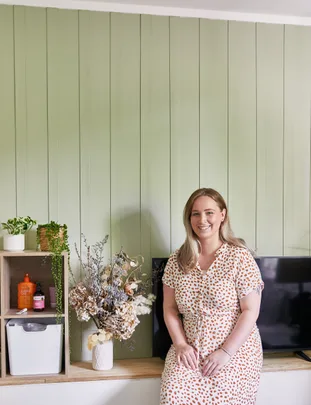 Woman in polka dot dress sitting on a bench, beside flowers and shelves, with green paneled wall background.