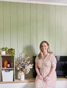 Woman in polka dot dress sitting on a bench, beside flowers and shelves, with green paneled wall background.