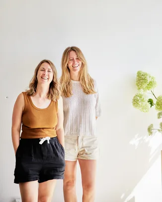 Two women standing and smiling in casual summer attire against a white wall with green floral decor.