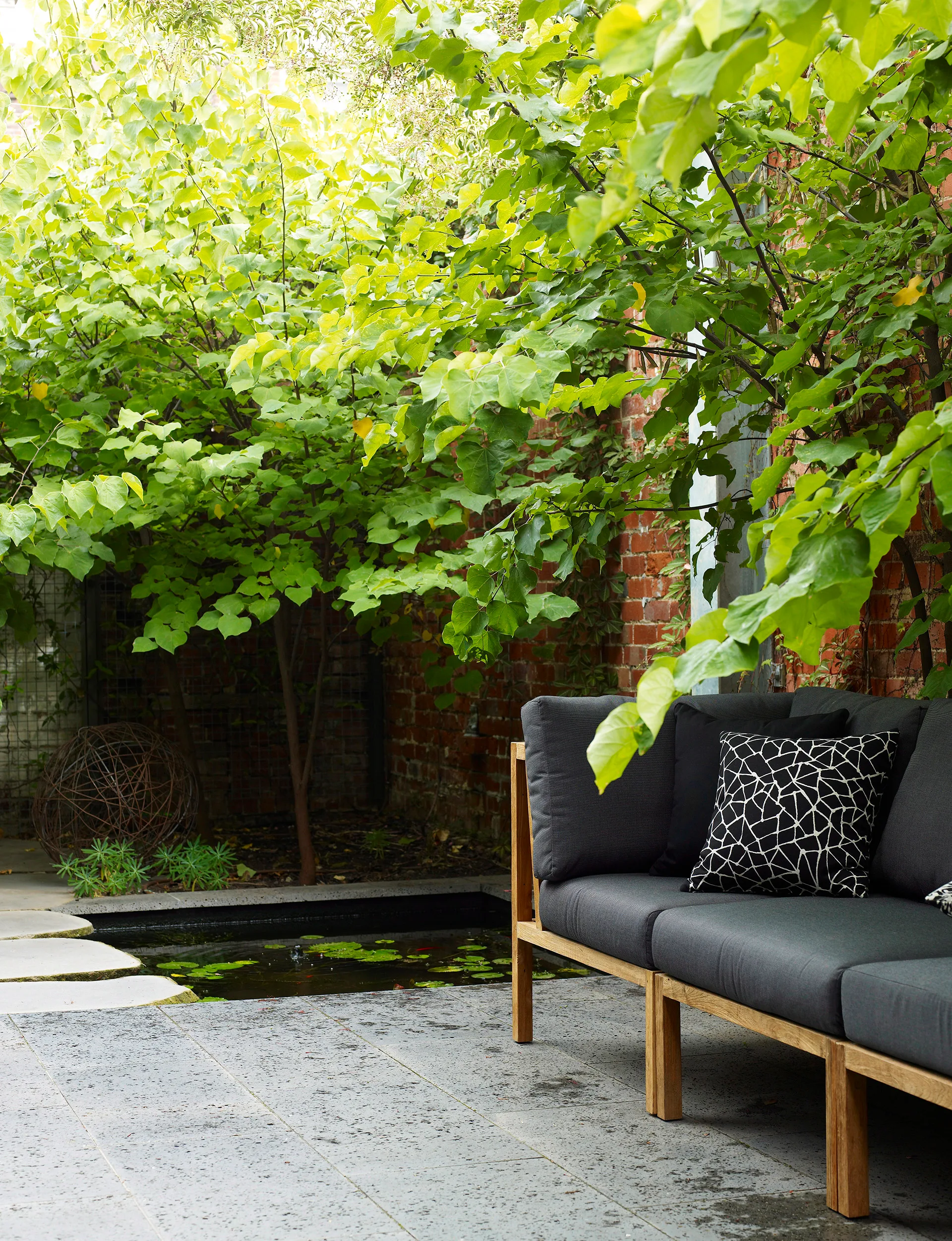 Outdoor seating area with gray cushioned bench, geometric pillow, and leafy green trees beside a small pond.