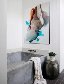 Concrete bathtub with a white towel, abstract painting on wall, and small plant on a metallic side table.