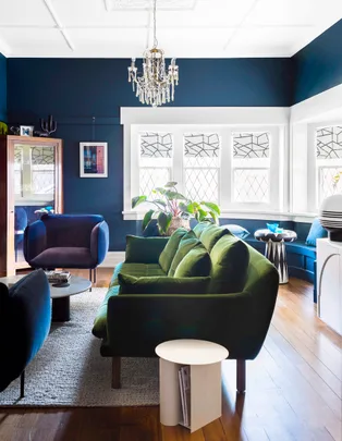 Living room with a green velvet sofa, blue armchairs, chandelier, and plants, featuring dark blue walls and wooden floor.
