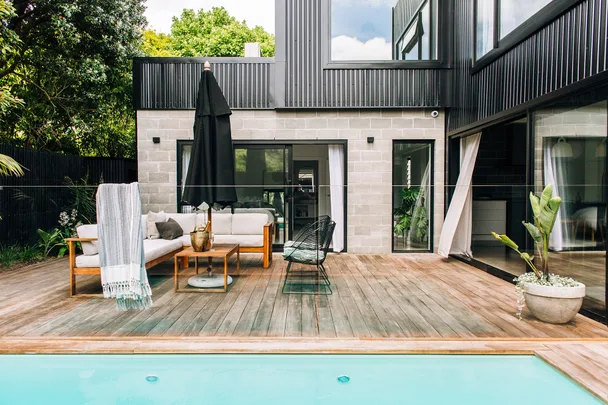 Modern patio with a pool, wooden deck, white sofa, umbrella, and plants against a black and gray house exterior.