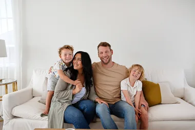 Family of four smiling and sitting on a white sofa in a living room setting.