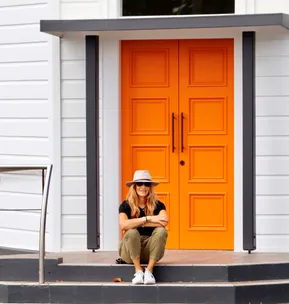 A person sits on steps in front of a bright orange door, wearing a hat, sunglasses, and casual attire.