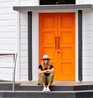 A person sits on steps in front of a bright orange door, wearing a hat, sunglasses, and casual attire.
