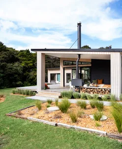 Modern house with wood-paneled exterior, large windows, and porch with seating area set in a lush green landscape.