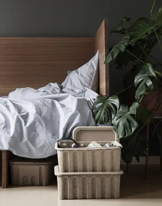 Bedroom with wooden headboard, white bedding, Monstera plant, and two stacked beige storage boxes.