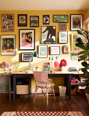 A home office with a desk, chair, and wall adorned with colorful framed artwork on a yellow background.