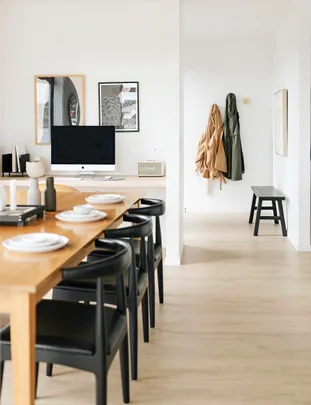 Modern dining area with a wooden table, black chairs, computer screen, and coat hangers on a light-colored wall.