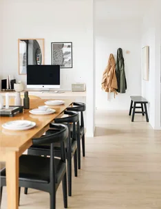 Modern dining area with a wooden table, black chairs, computer screen, and coat hangers on a light-colored wall.
