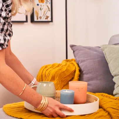 Person placing a tray with candles and a cup on a yellow blanket, next to colorful cushions.