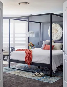 Four-poster bed in modern bedroom with white bedding, orange throw, decorative pillows, and slippers on a patterned rug.