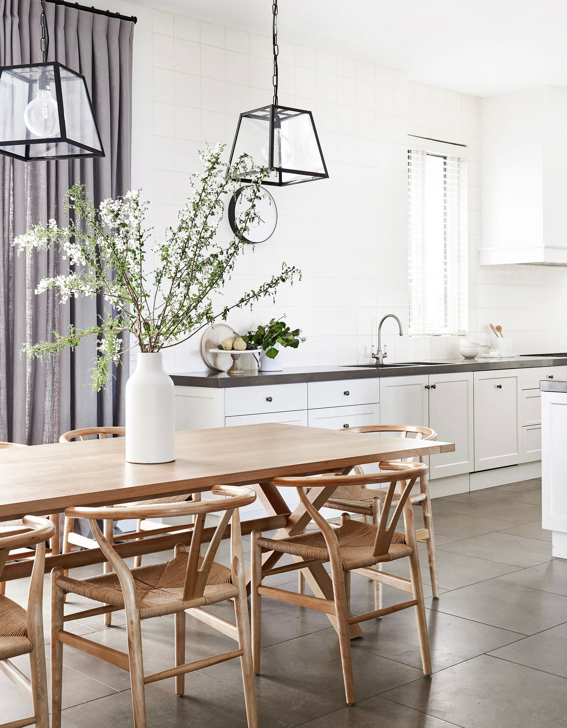Modern kitchen with wooden dining set, large vase of white flowers, white cabinets, and geometric pendant lights.