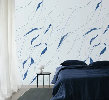 Bedroom with a navy blue bed, abstract blue leaf-patterned wall, sheer curtains, and a small side table with a white vase.