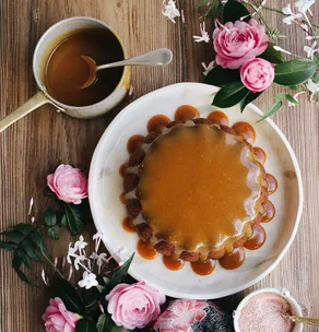 Caramel-glazed cake on a white plate with roses, a saucepan of caramel, and a bowl of pink salt on a wooden table.