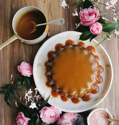 Caramel-glazed cake on a white plate with roses, a saucepan of caramel, and a bowl of pink salt on a wooden table.