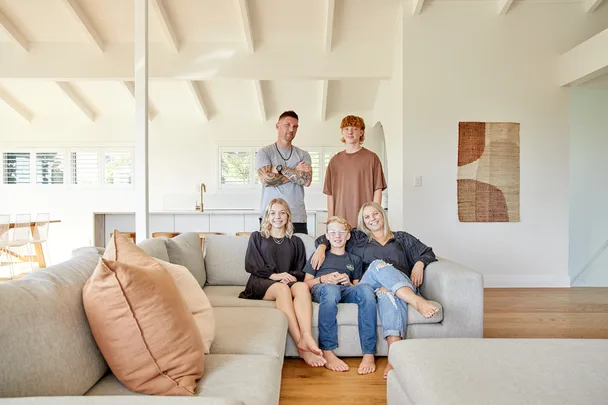 Family of five sitting on a large couch in a bright, modern living room with wooden floors and minimal decor.