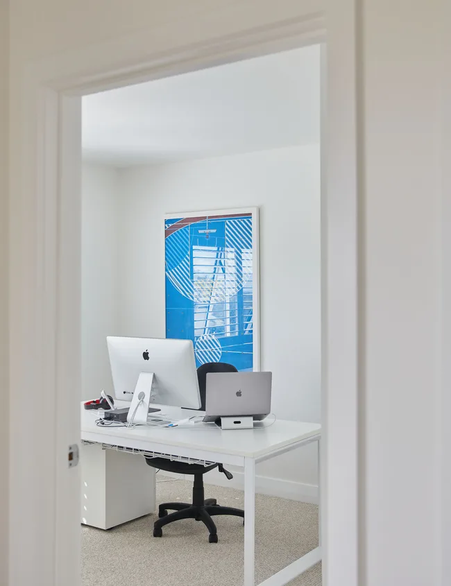 Modern home office with iMac, laptop, and abstract blue art on the wall, viewed through a doorframe.