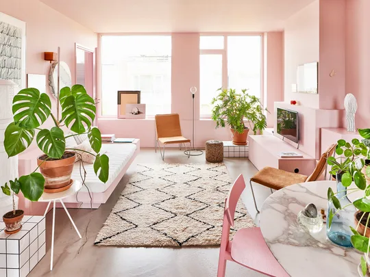 Bright, modern living room with pink walls, green plants, white furniture, and geometric rug pattern.