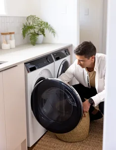 Man doing laundry, placing wicker basket near white washing machine in a modern laundry room with a plant on the counter.