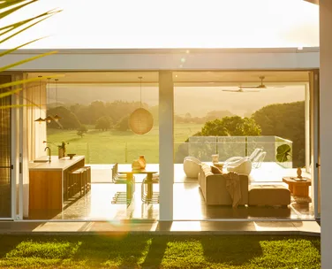 Modern open-plan living and dining area with glass walls, overlooking a green landscape in bright sunlight.