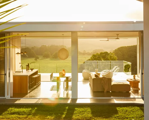 Modern open-plan living and dining area with glass walls, overlooking a green landscape in bright sunlight.