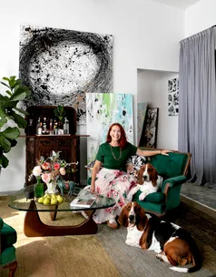 A woman sits on a green sofa with two dogs in an art-filled living room with abstract paintings on the wall.
