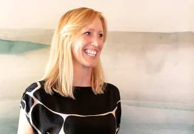 A woman with blonde hair smiling, wearing a black and white patterned blouse, stands against a blurred background.