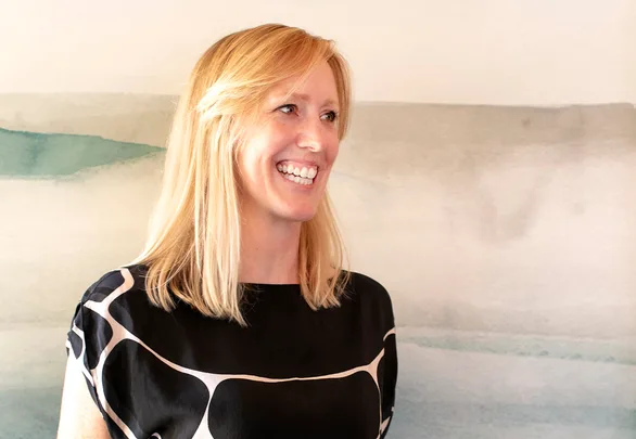 A woman with blonde hair smiling, wearing a black and white patterned blouse, stands against a blurred background.