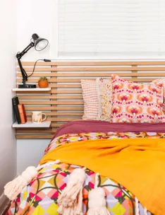 Cozy bedroom with colorful bedding, patterned pillows, bedside lamp, books, and a mug on a shelf.