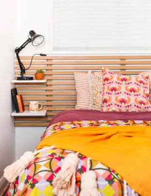 Cozy bedroom with colorful bedding, patterned pillows, bedside lamp, books, and a mug on a shelf.