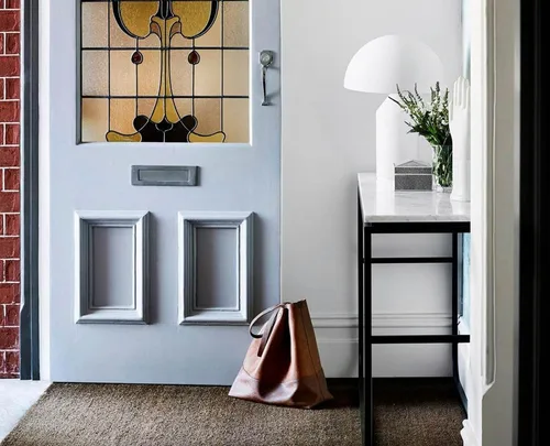 Stained glass door with a brown bag on the floor, a white table with a plant and lamp in a home's entryway.